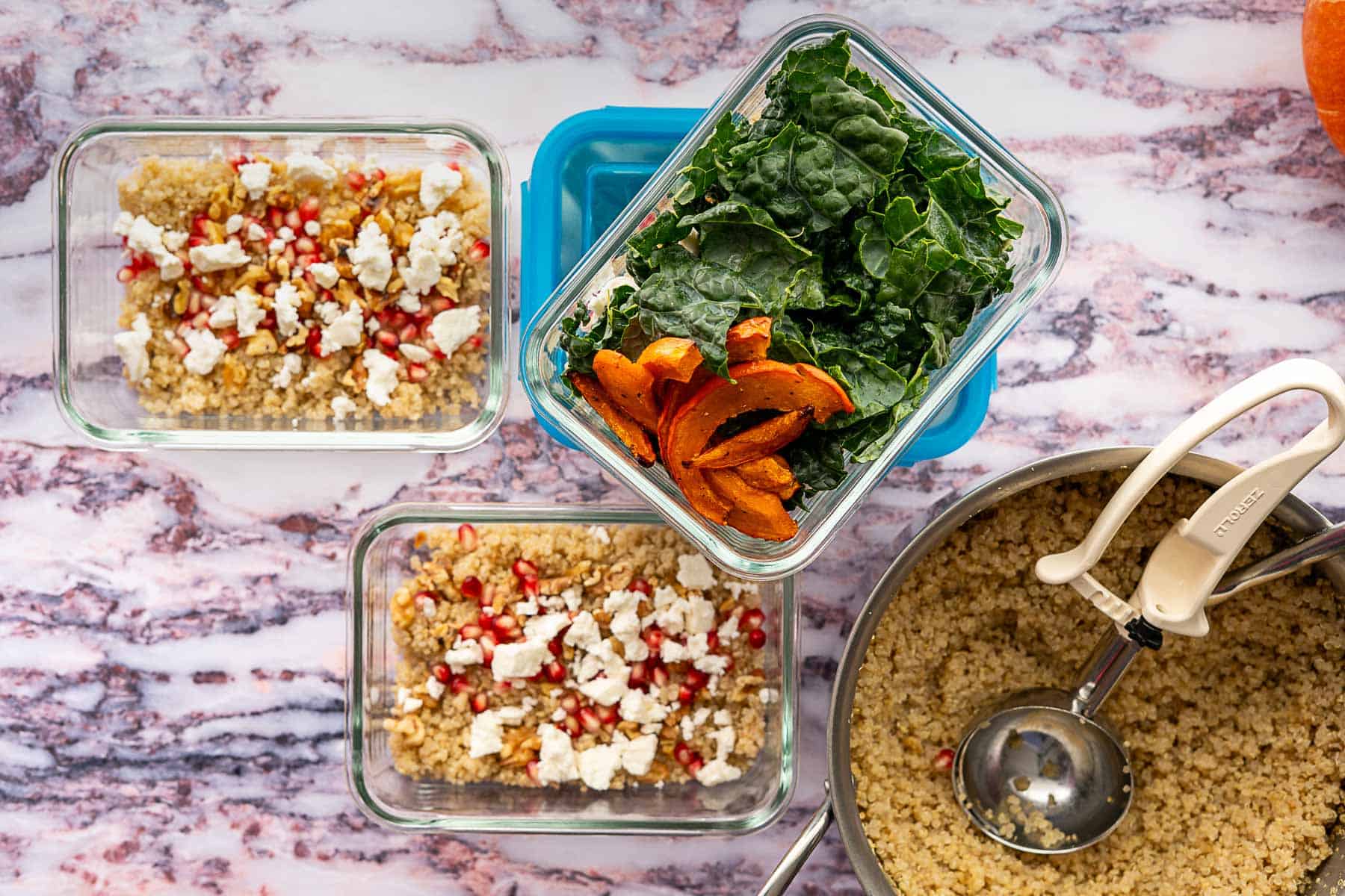 Two glass containers with quinoa, crumbled feta, and pomegranate seeds sit beside a container of kale and roasted squash—a colorful kale and pomegranate salad. Nearby, a pot of cooked quinoa with a ladle rests on a marble surface.