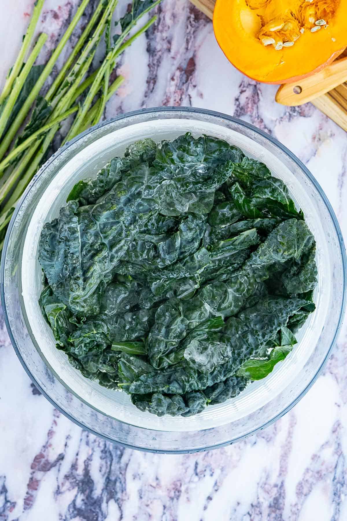 A glass bowl filled with dark green kale leaves sits on a marble surface, ready for a fresh kale and pomegranate salad. Nearby, raw green onions and a partially cut yellow squash rest on a wooden cutting board.