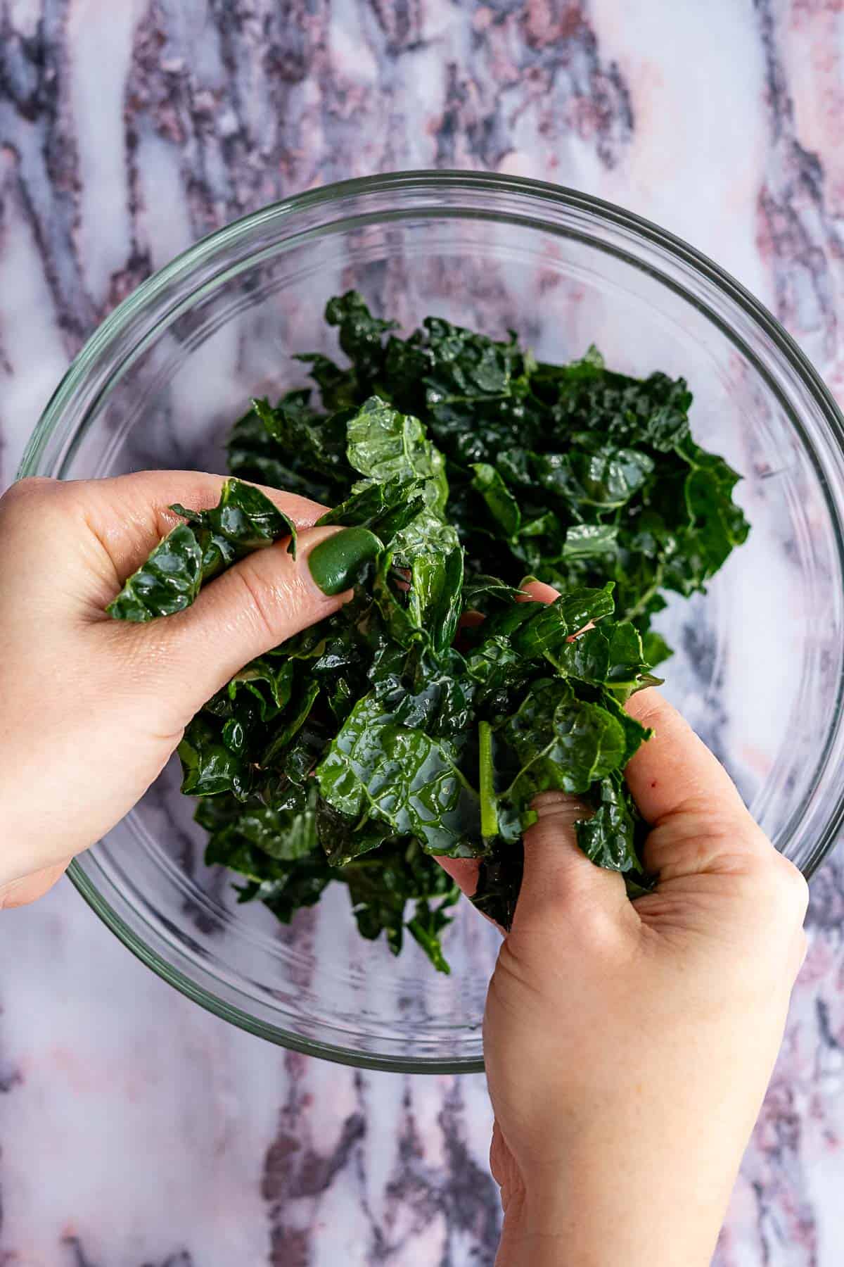 Hands massaging chopped kale leaves in a clear glass bowl on a marble countertop, preparing the base for a vibrant kale and pomegranate salad.
