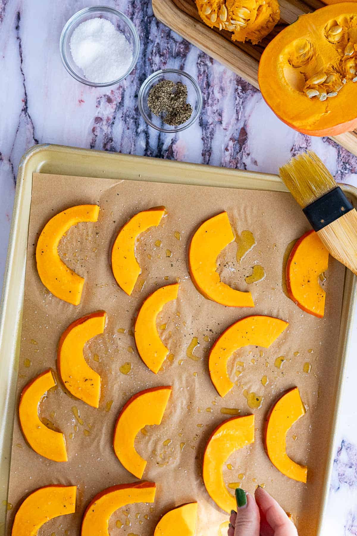 Sliced red kuri squash arranged on a parchment-lined baking sheet, drizzled with oil. Nearby are bowls of salt and pepper, a brush, and a halved squash on a cutting board, perfect for pairing with kale and pomegranate salad. A hand is sprinkling salt.