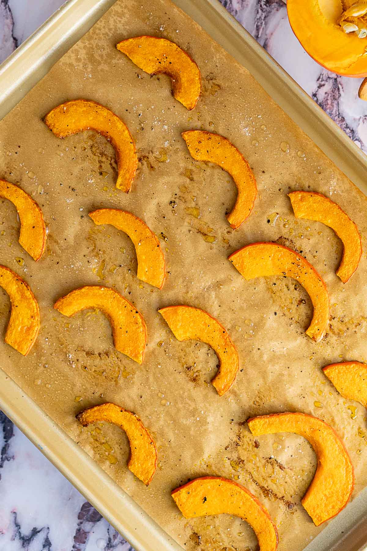 Sliced roasted red kuri squash pieces arranged on a parchment-lined baking sheet, lightly seasoned and baked until golden, with a vibrant kale and pomegranate salad visible in the corner on a marble surface.