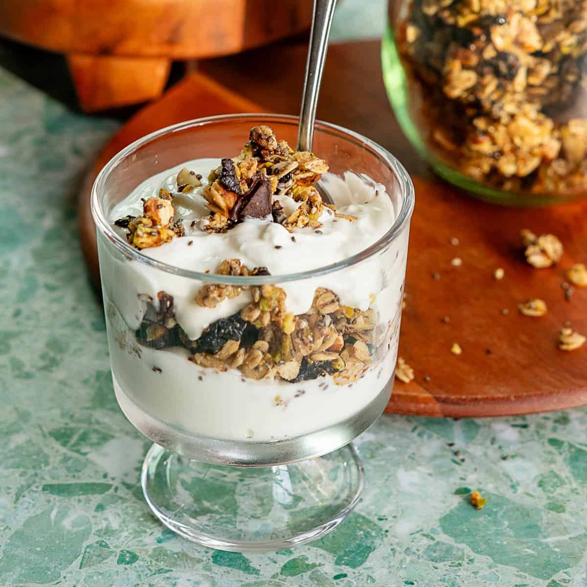A glass cup filled with layers of creamy yogurt and granola, topped with more granola and a spoon, sits on a green marble surface. A jar of granola and scattered crumbs are visible in the background.