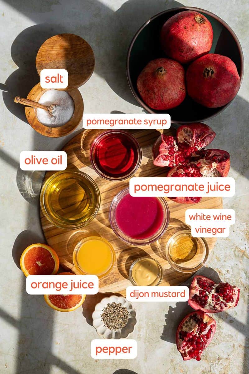 A top view of ingredients for a pomegranate vinaigrette on a light surface, including salt, olive oil, orange juice, pomegranate juice, pomegranate syrup, white wine vinegar, Dijon mustard, pepper, pomegranates, and an orange.
