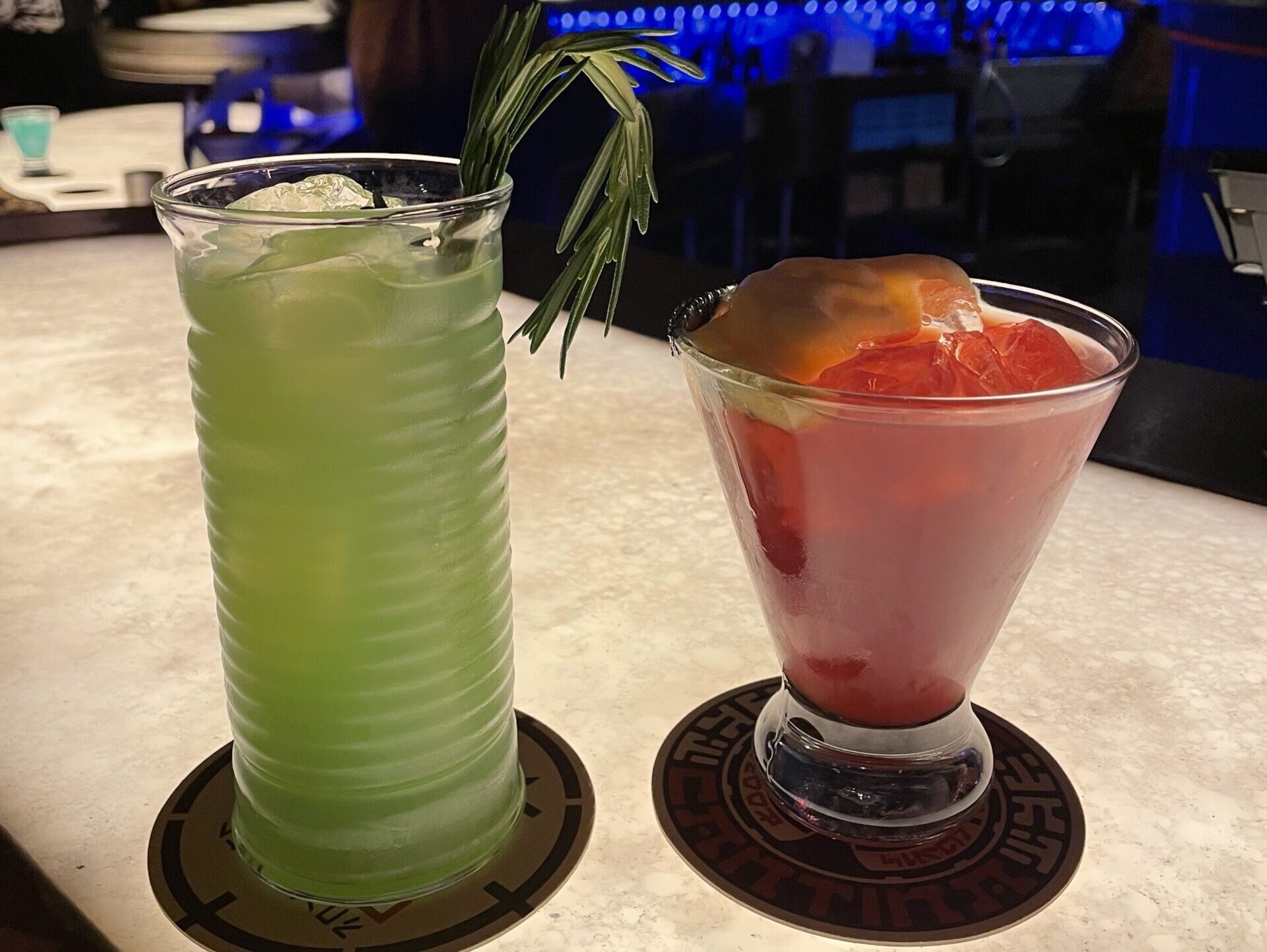 Two colorful cocktails sit on coasters; one is a green Dagobah Slug Slinger with ice and a rosemary garnish in a tall glass, the other is The Outer Rim Margarita in a short glass, both on a lit bar counter.