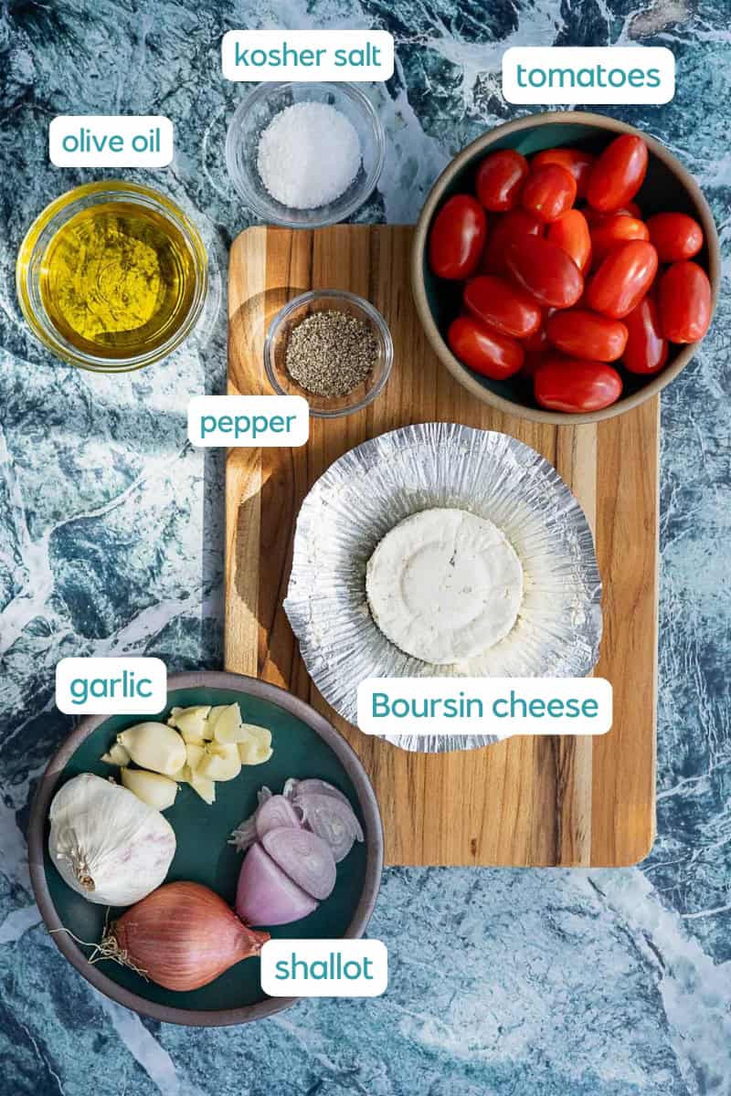 A top view of ingredients on a blue-marble surface: a bowl of grape tomatoes, a bowl of olive oil, a bowl of kosher salt, ground pepper, Boursin cheese, garlic cloves, and a shallot on a cutting board. Labels identify each item.