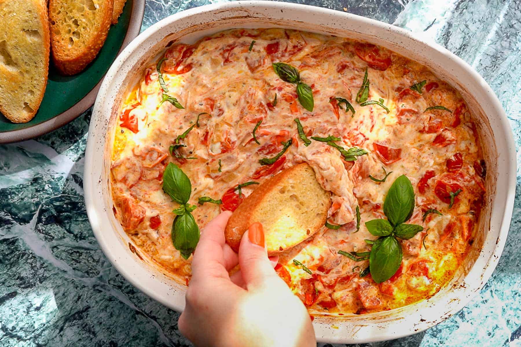 A hand dips a slice of toasted bread into a baked cheesy Boursin tomato dip in a white casserole dish, garnished with fresh basil leaves. Toasted bread slices are on a plate nearby.