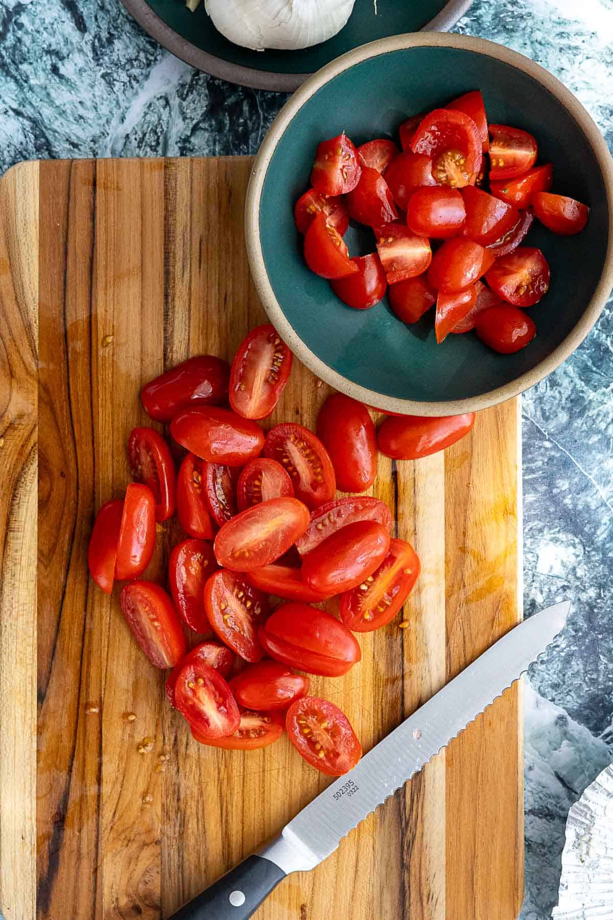 A wooden cutting board with halved grape tomatoes, a serrated knife, and a bowl filled with more cut tomatoes, set on a marble countertop.