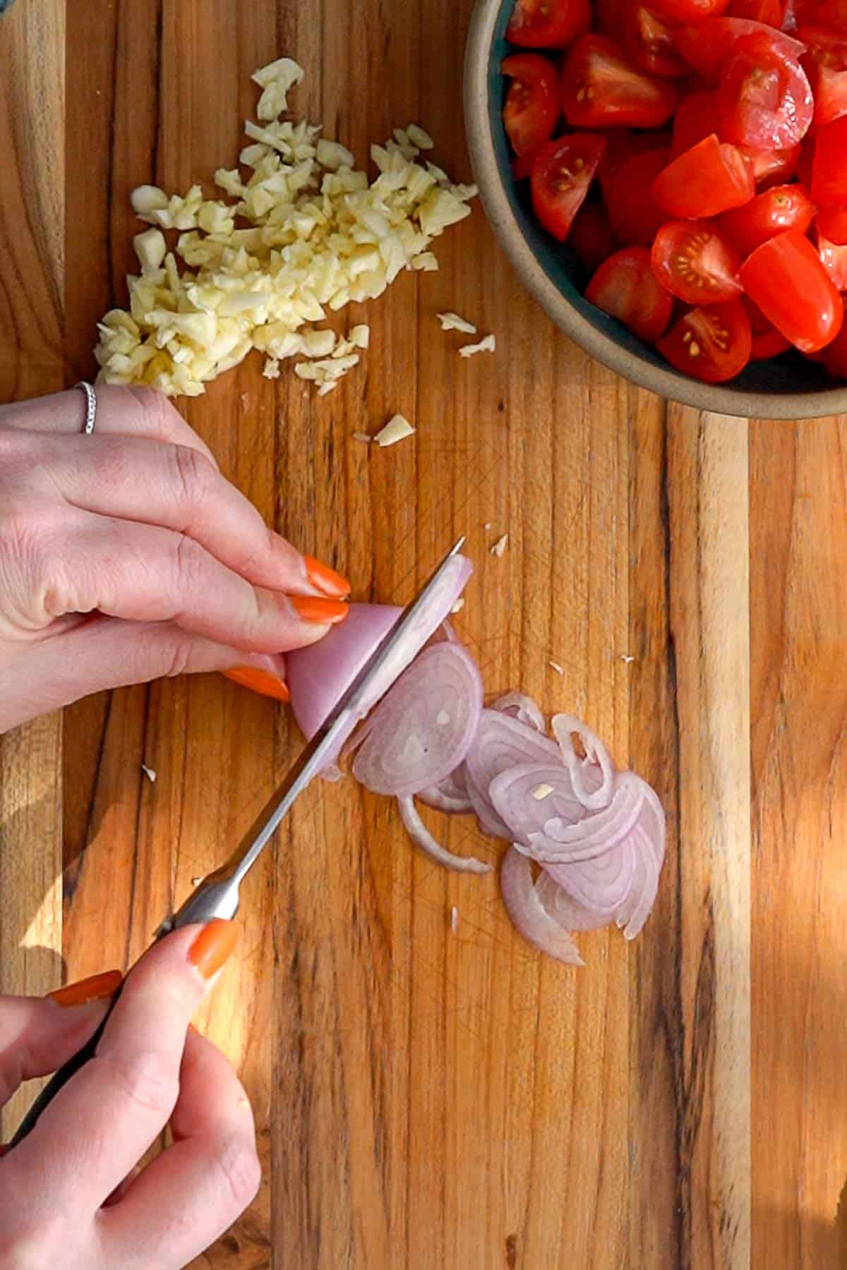 A person slices a shallot on a wooden cutting board with chopped garlic beside it. A bowl of halved cherry tomatoes is in the upper right corner.