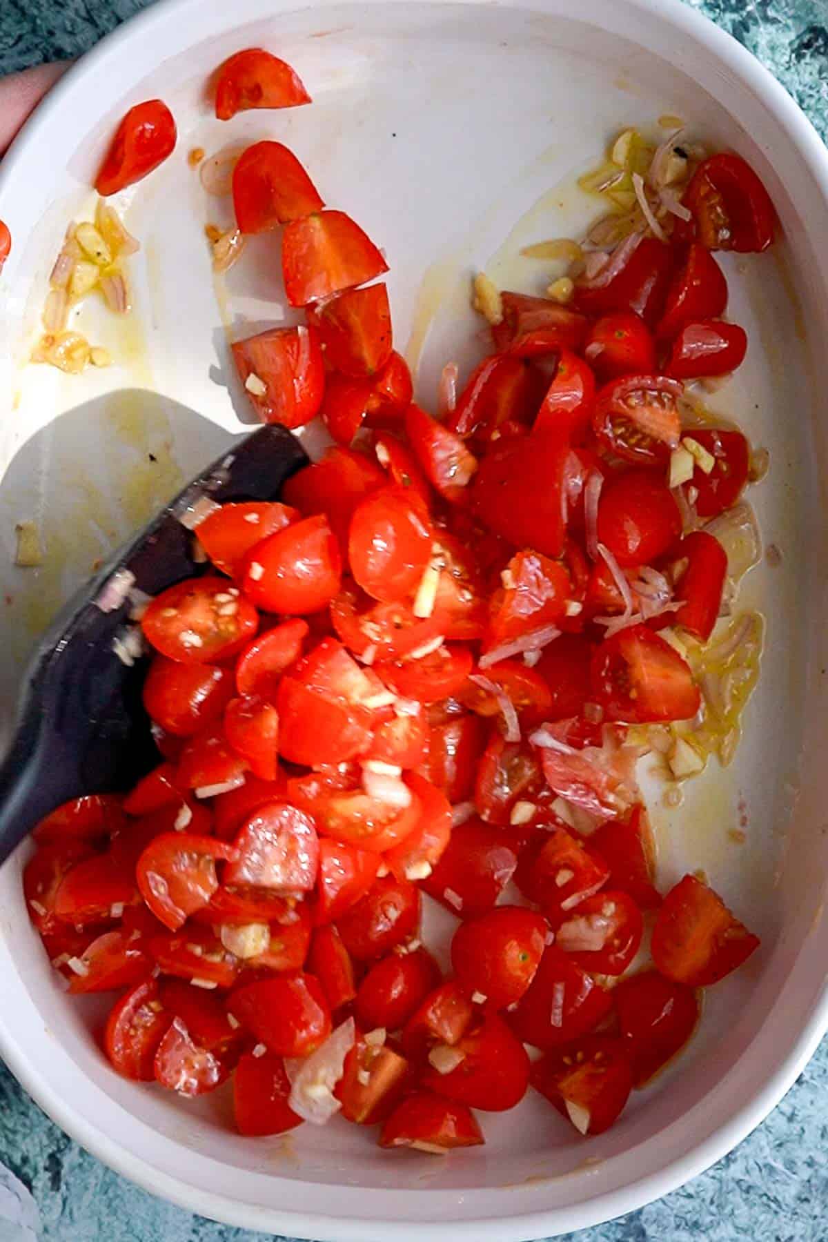 Chopped cherry tomatoes, minced garlic, and shallots are being stirred together in a white baking dish with a black spatula.