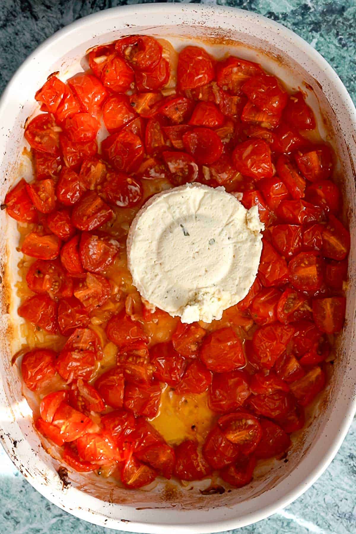 A white baking dish filled with roasted cherry tomatoes surrounding a round block of creamy white Boursin cheese, placed on a green marbled countertop.