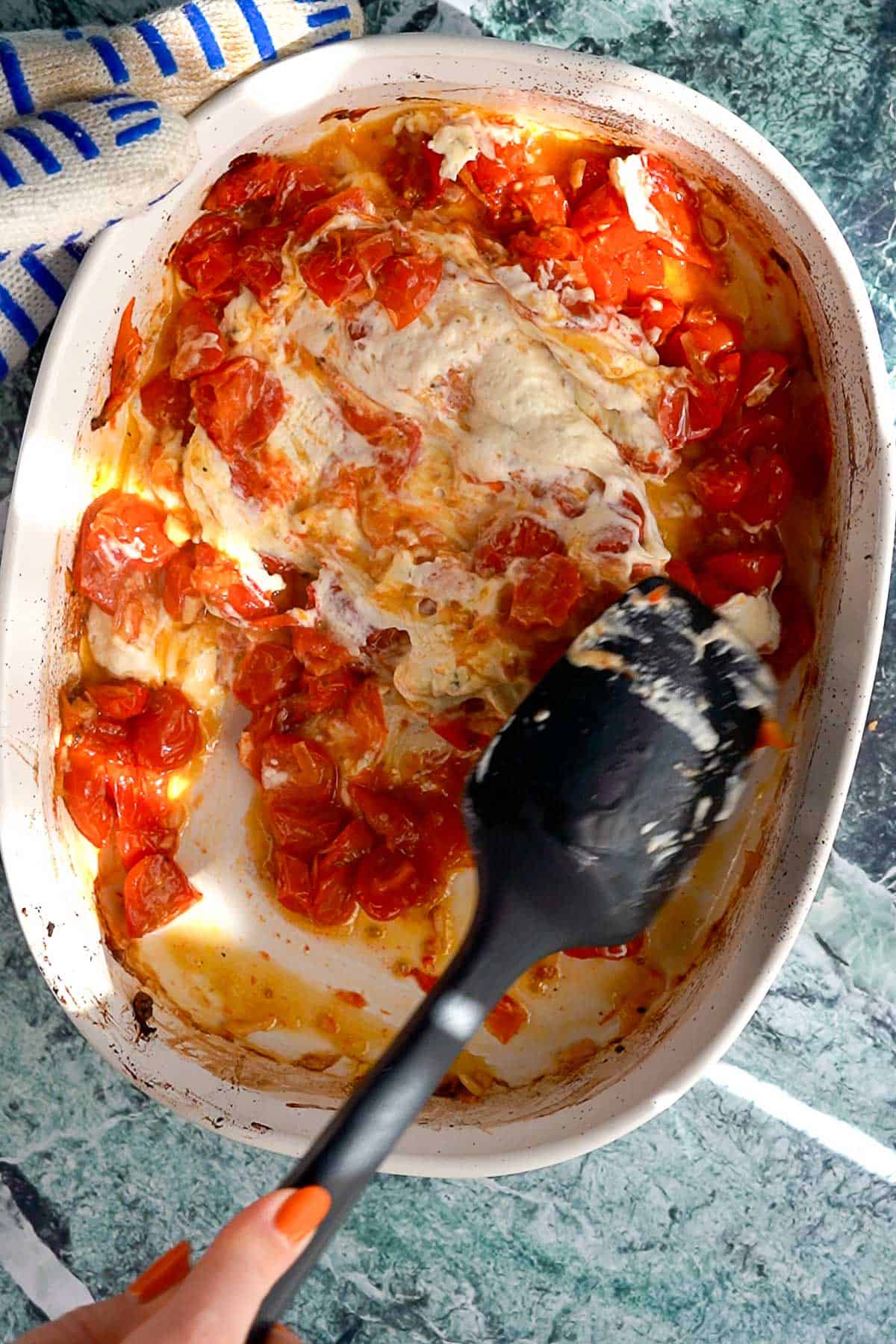 A hand holding a black spatula mixes melted cheese and roasted cherry tomatoes in a white baking dish on a green countertop. A blue and white kitchen towel is nearby.