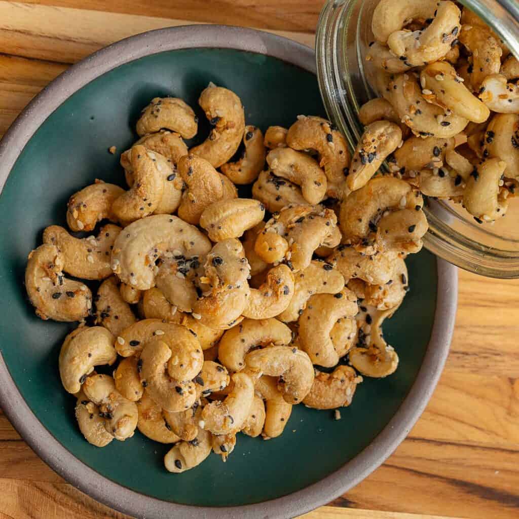 A green ceramic bowl filled with everything bagel cashews coated in black and white sesame seeds, with more cashews spilling from a glass jar onto the bowl, all arranged on a wooden surface.