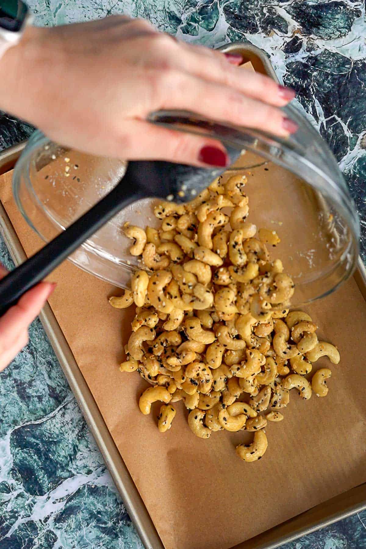 A person pours everything bagel cashews from a glass bowl onto a parchment-lined baking sheet, using a black spatula, over a green and white marbled countertop.