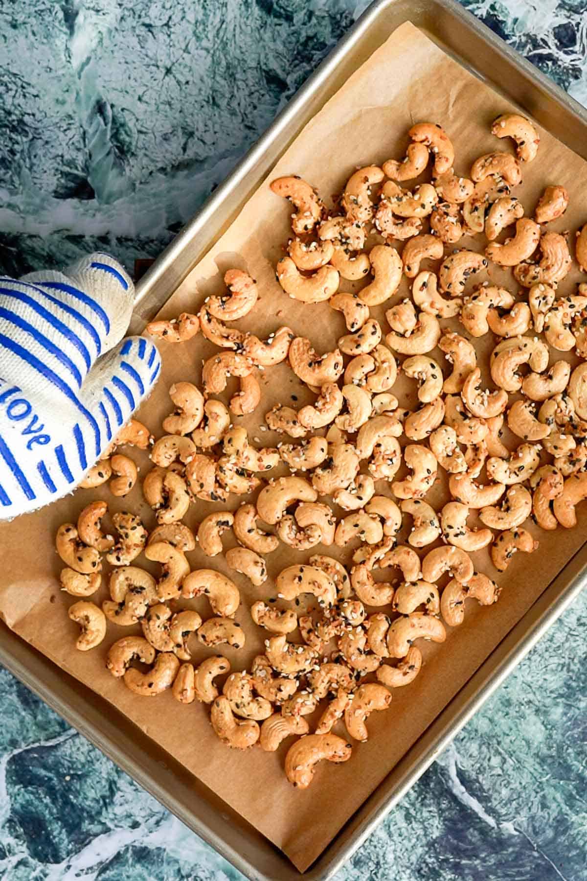 A hand wearing a blue and white oven mitt holds a baking tray lined with parchment paper, topped with everything bagel cashews sprinkled with seeds and seasoning, on a green marble countertop.