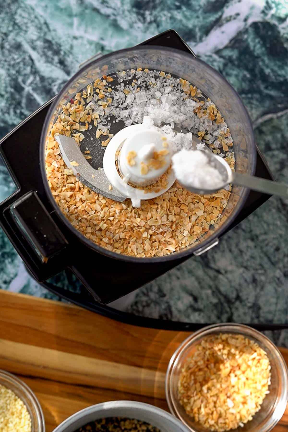 A food processor filled with dried onion flakes and salt, with a spoon adding more salt, sits on a countertop next to small bowls of additional ingredients.