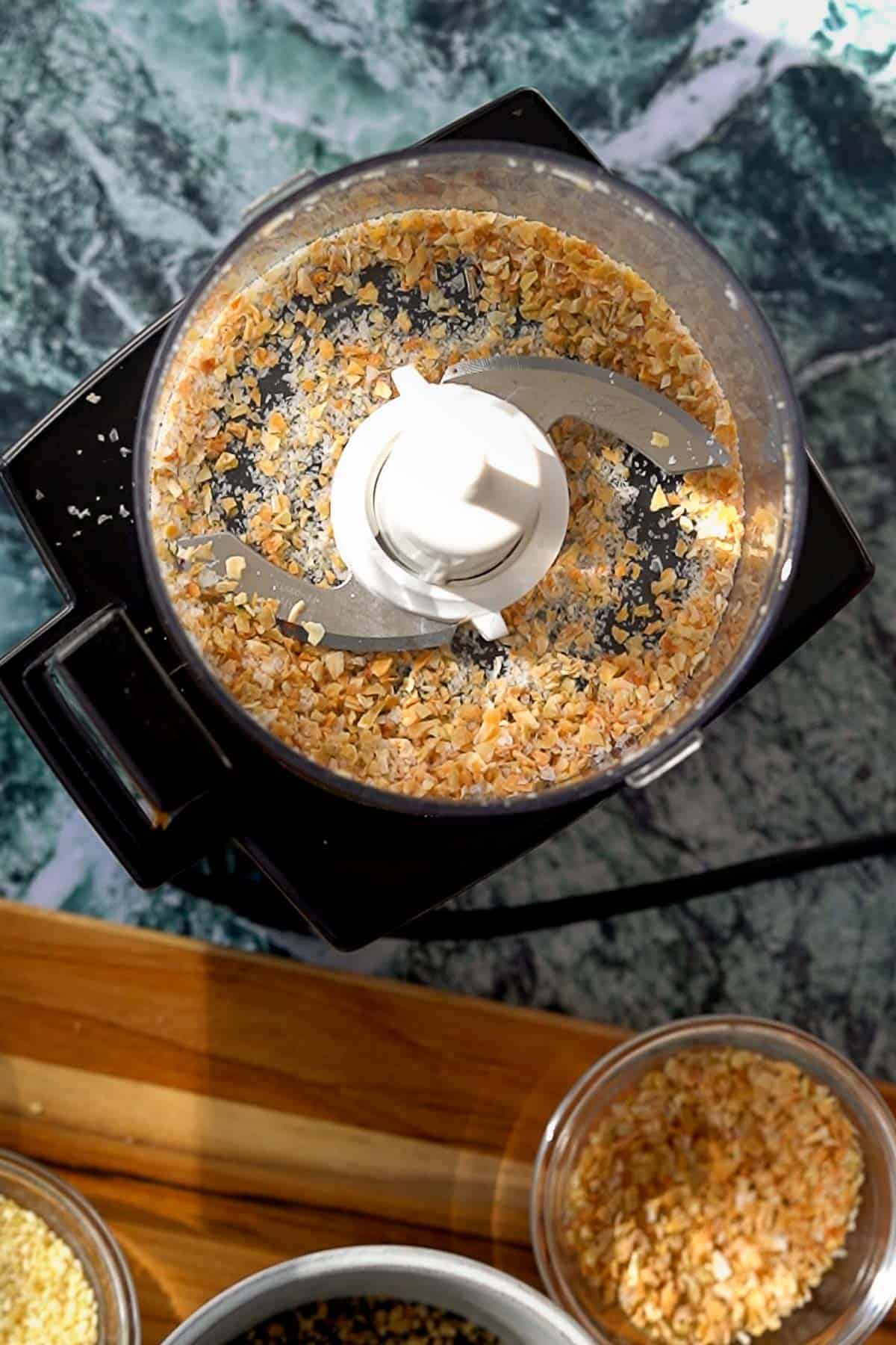 A food processor with a metal blade contains coarsely ground dried onions. Nearby, bowls of additional dried onion pieces sit on a wooden surface. The background features a marbled green and white countertop.