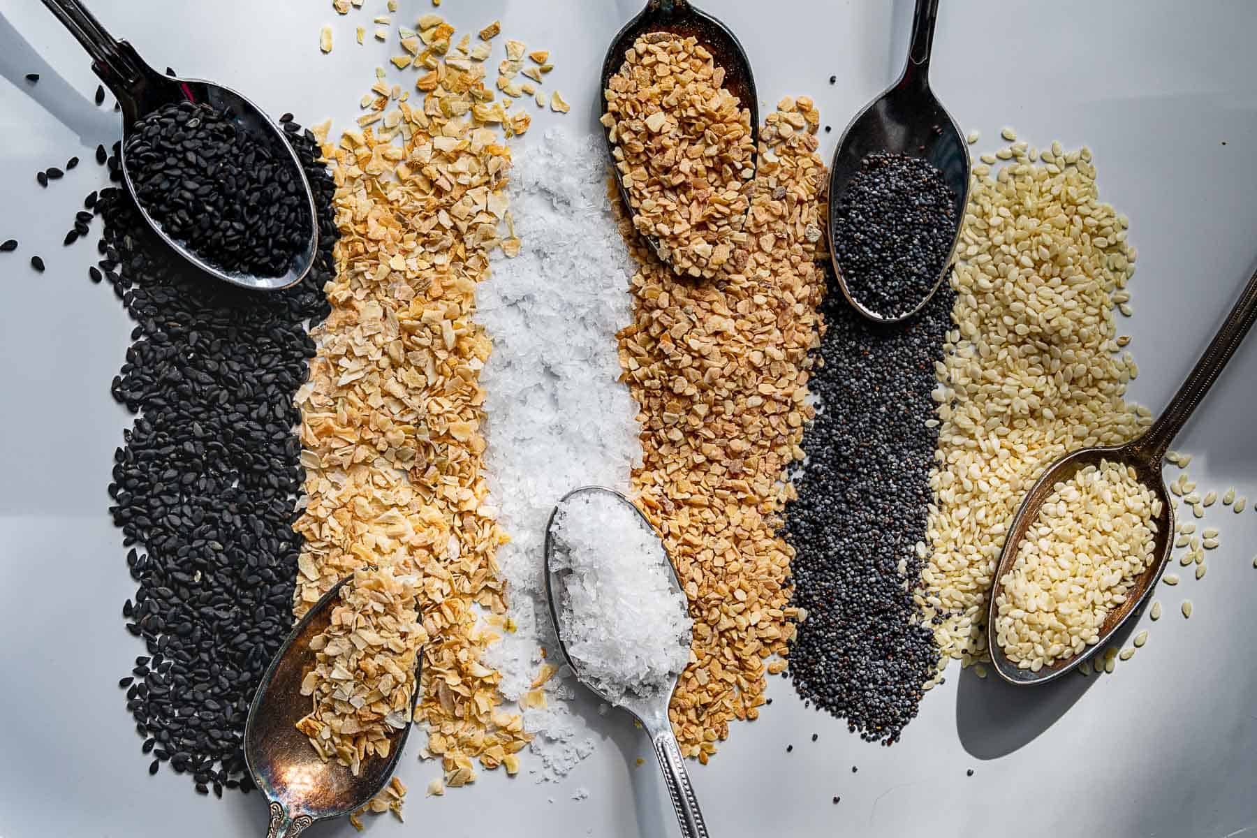 Five metal spoons hold black sesame seeds, rolled oats, brown sugar, poppy seeds, and white sesame seeds, each forming a stripe of spilled contents on a white surface.