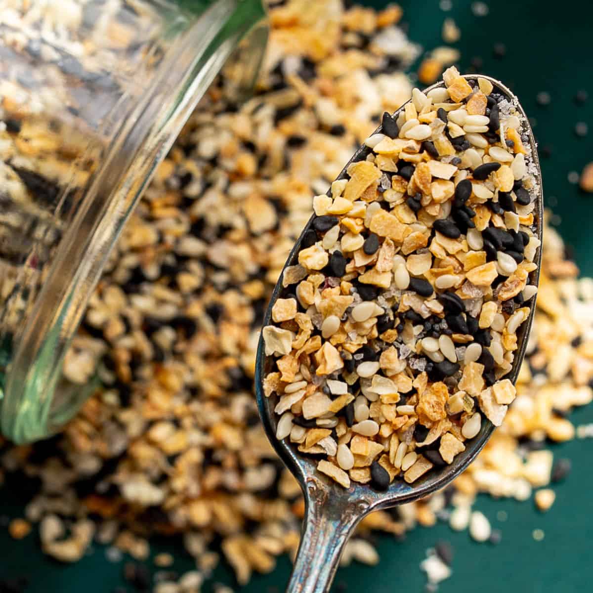 A spoonful of everything bagel seasoning, with sesame seeds, dried garlic, dried onion, and black sesame seeds, held above a green surface with more seasoning spilled from a glass jar in the background.
