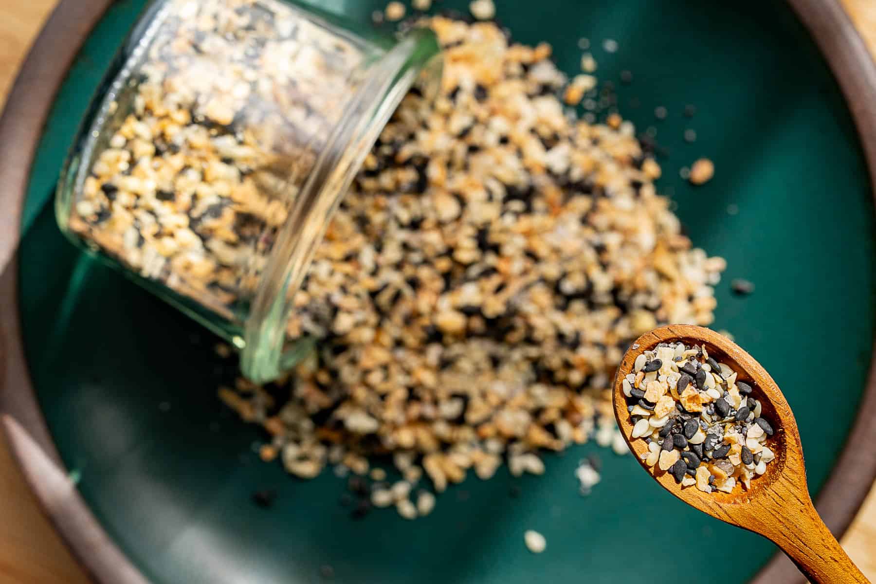 A glass jar tipped over on a dark green plate, spilling homemade everything bagel seasoning, a mixture of black and white seeds with dried herbs. A wooden spoon holds a scoop of the mixture in the foreground.