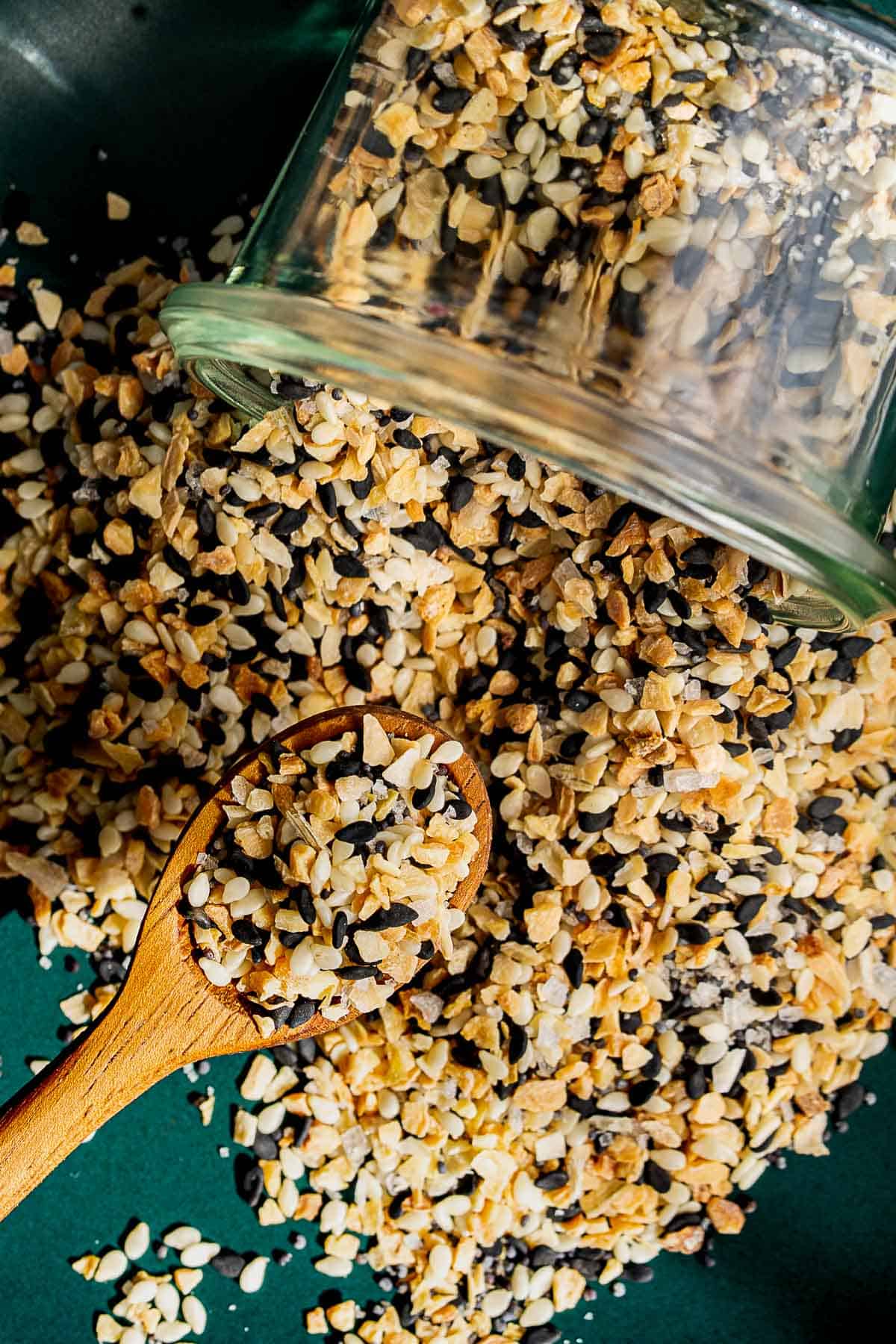 A glass jar of everything bagel seasoning spills onto a green surface, with a wooden spoon scooping up the mixture of sesame seeds, poppy seeds, dried garlic, dried onion, and salt.