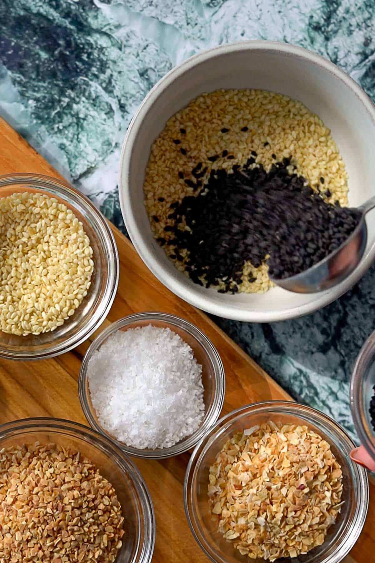 A spoon adds black sesame seeds to a bowl of white sesame seeds. Surrounding the bowl are small glass bowls filled with more sesame seeds, flaky salt, dried garlic, and dried onion on a wooden surface.