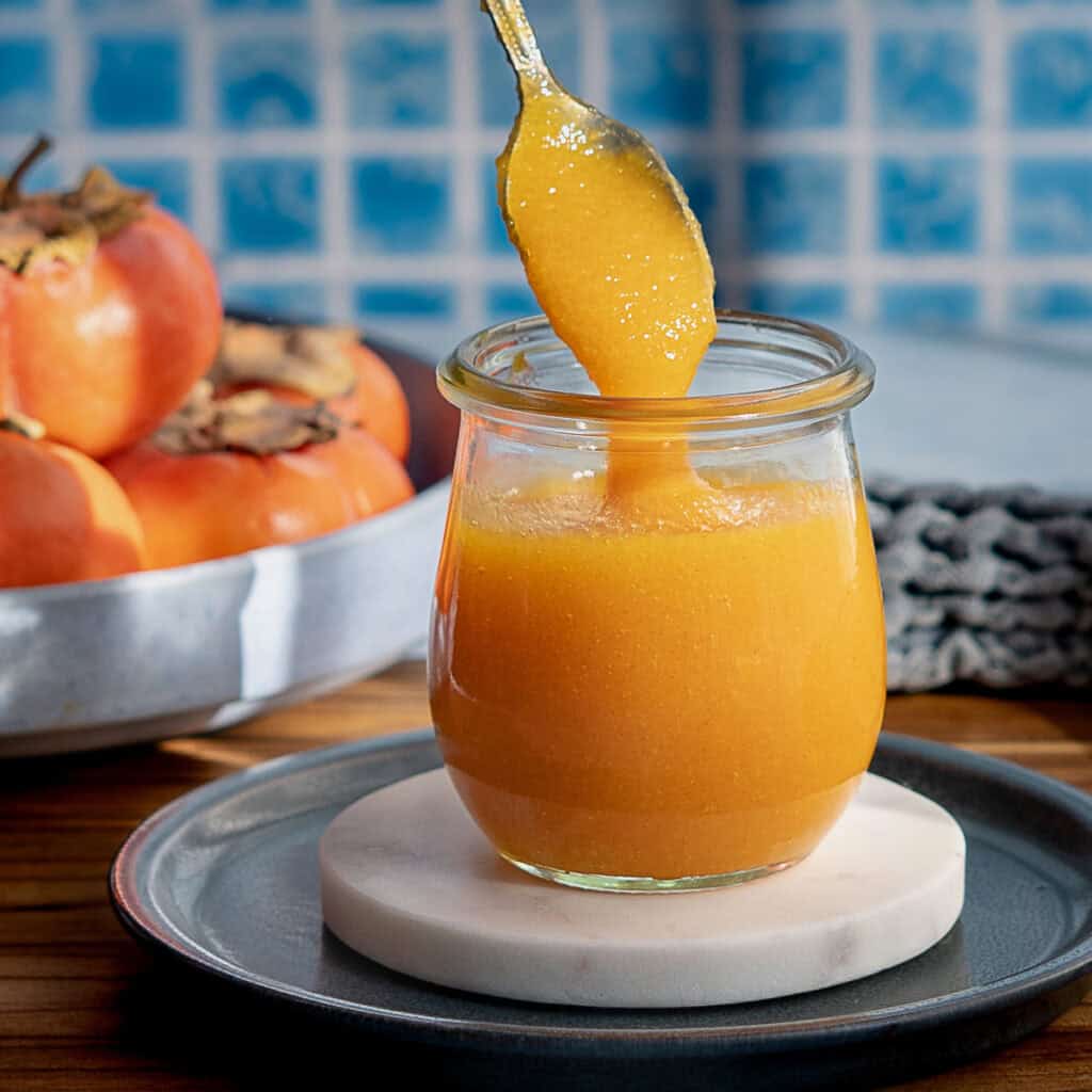 A spoon lifts thick, orange persimmon jam from a glass jar placed on a marble coaster and gray plate. Fresh persimmons sit in a bowl in the background against a blue tile wall.