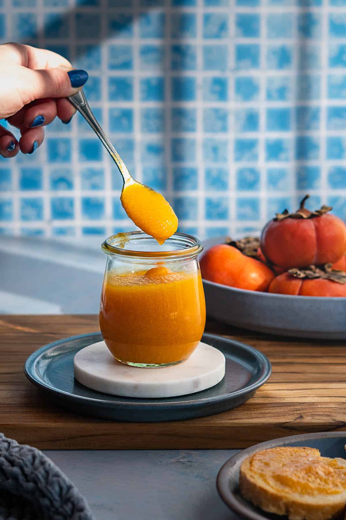 A hand holds a spoonful of persimmon jam above a small glass jar. In the background, a bowl of whole persimmons sits on the table, while in the foreground, a slice of bread spread with sweet persimmon jam is visible.