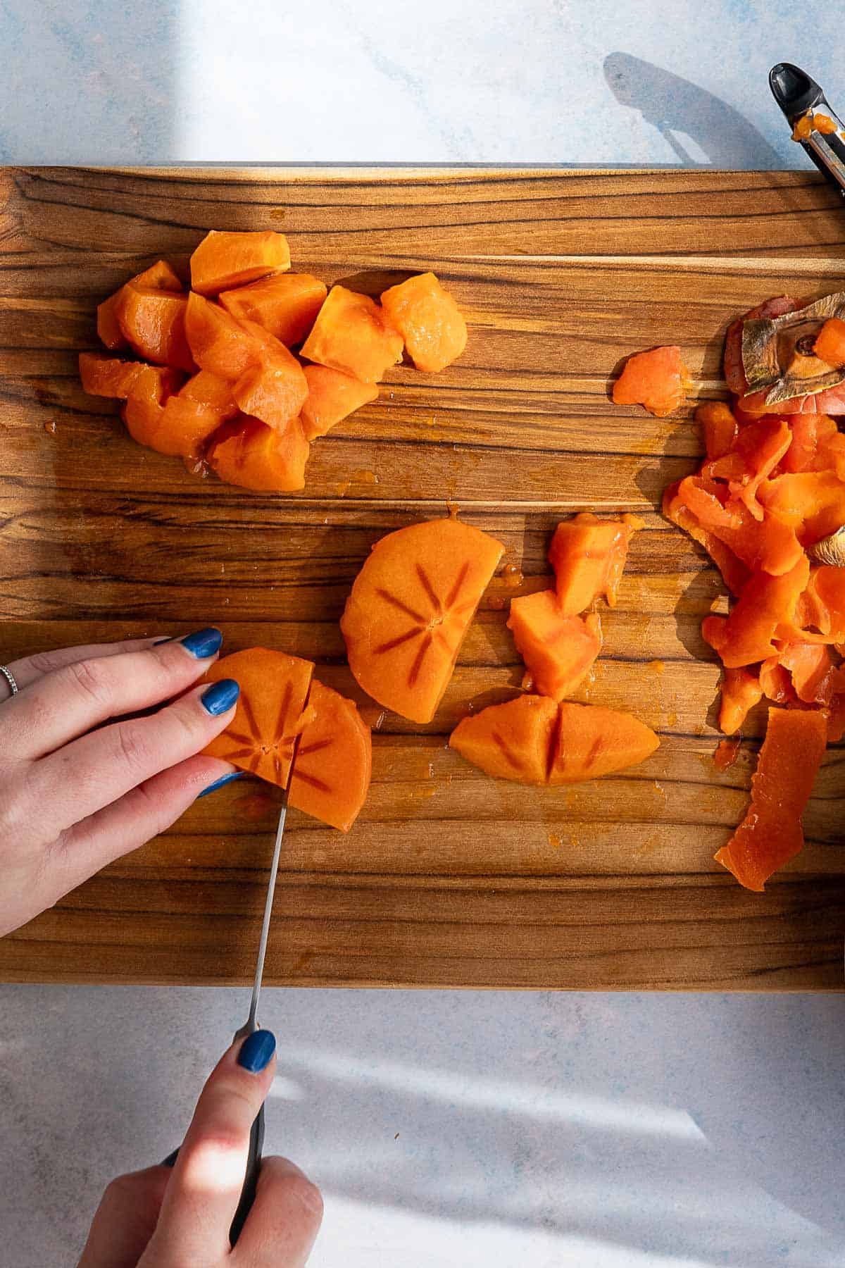 A person with blue nail polish slices orange persimmon fruit—perfect for making persimmon jam—on a striped wooden cutting board, with diced pieces and peels nearby.