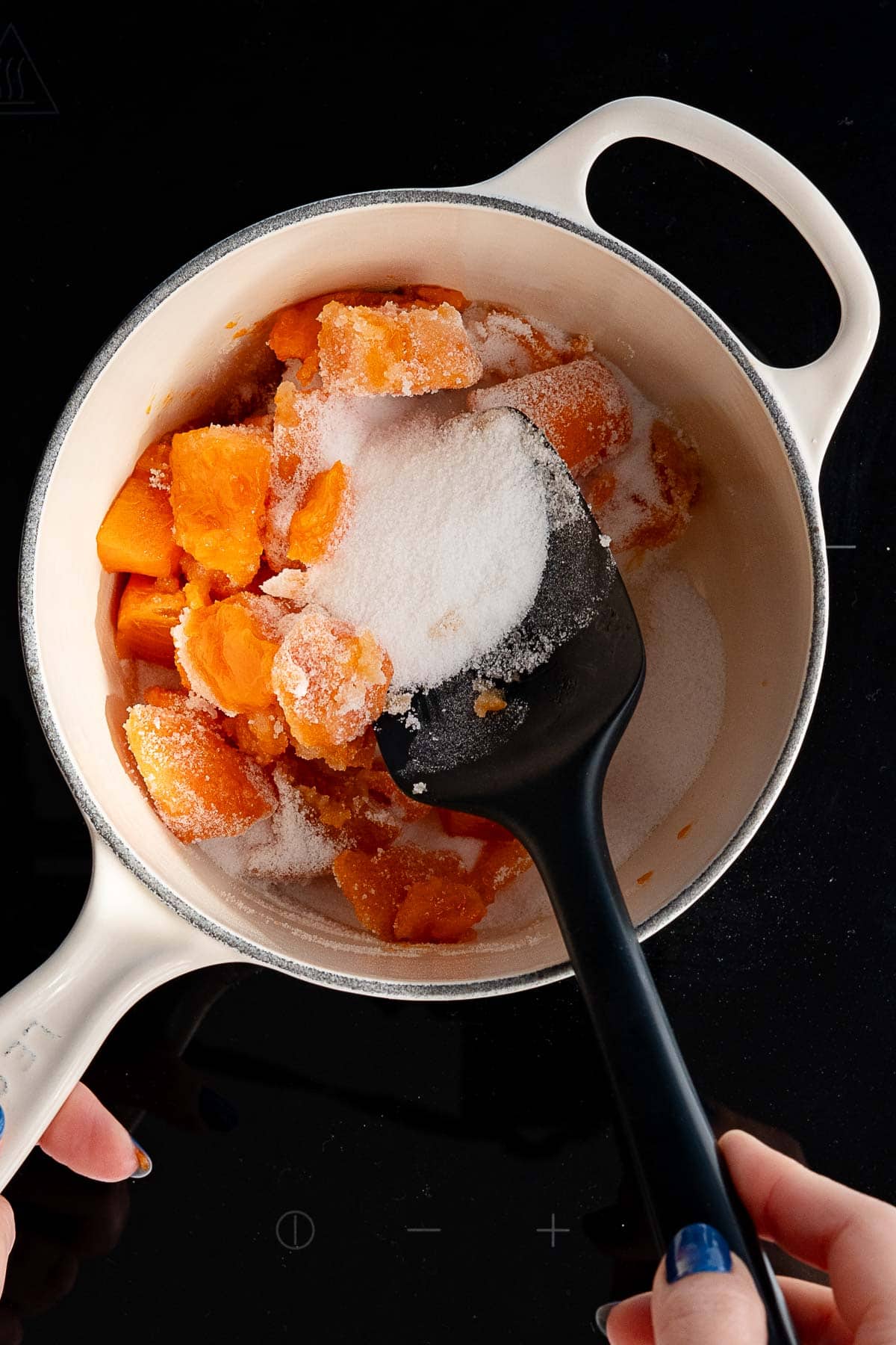 A black spatula stirs chopped orange fruit and sugar together for persimmon jam in a white pot, viewed from above, while a person holds the pot handle on a stovetop.