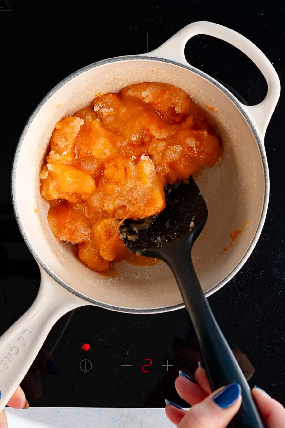 A hand stirs orange persimmon jam in a white pot on a stovetop, with the stove display showing the heat setting at 2.
