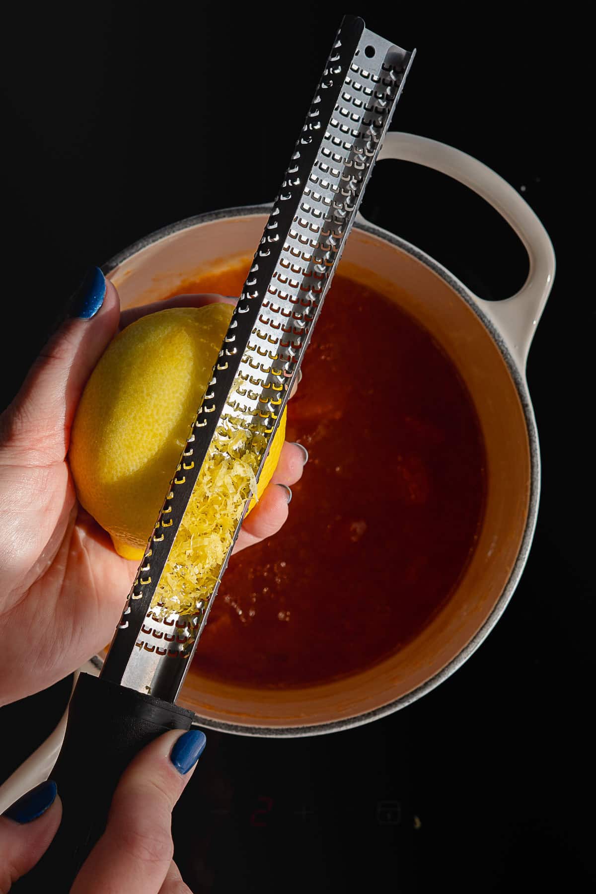 A hand grates lemon zest over a pot of reddish-orange persimmon jam on the stove, with the zest collecting on the metal grater. The person has blue nail polish.