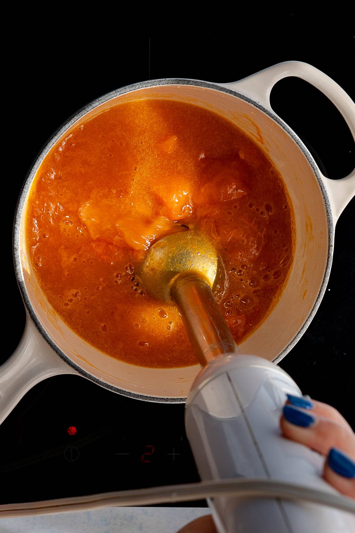 A person uses an immersion blender to puree a pot of orange soup or persimmon jam on a stovetop. The person's hand, with blue nail polish, holds the blender inside a light-colored pot.