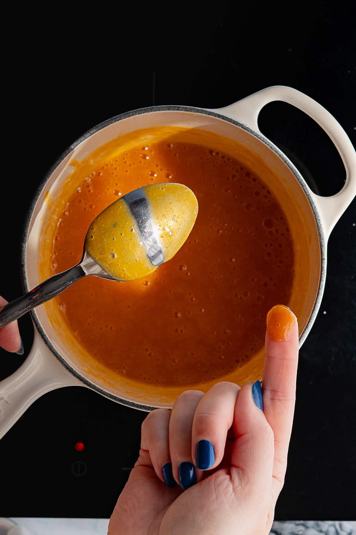A hand with blue nail polish holds a finger covered in orange persimmon jam, while another hand holds a spoon coated with the same jam over a pot filled with it.