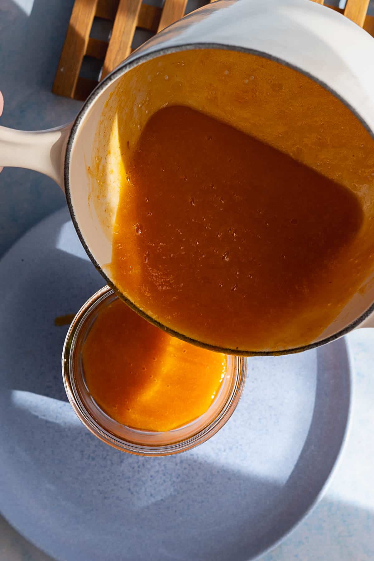 A hand pours thick orange persimmon jam from a white saucepan into a glass jar on a light blue plate. Sunlight casts shadows across the scene.