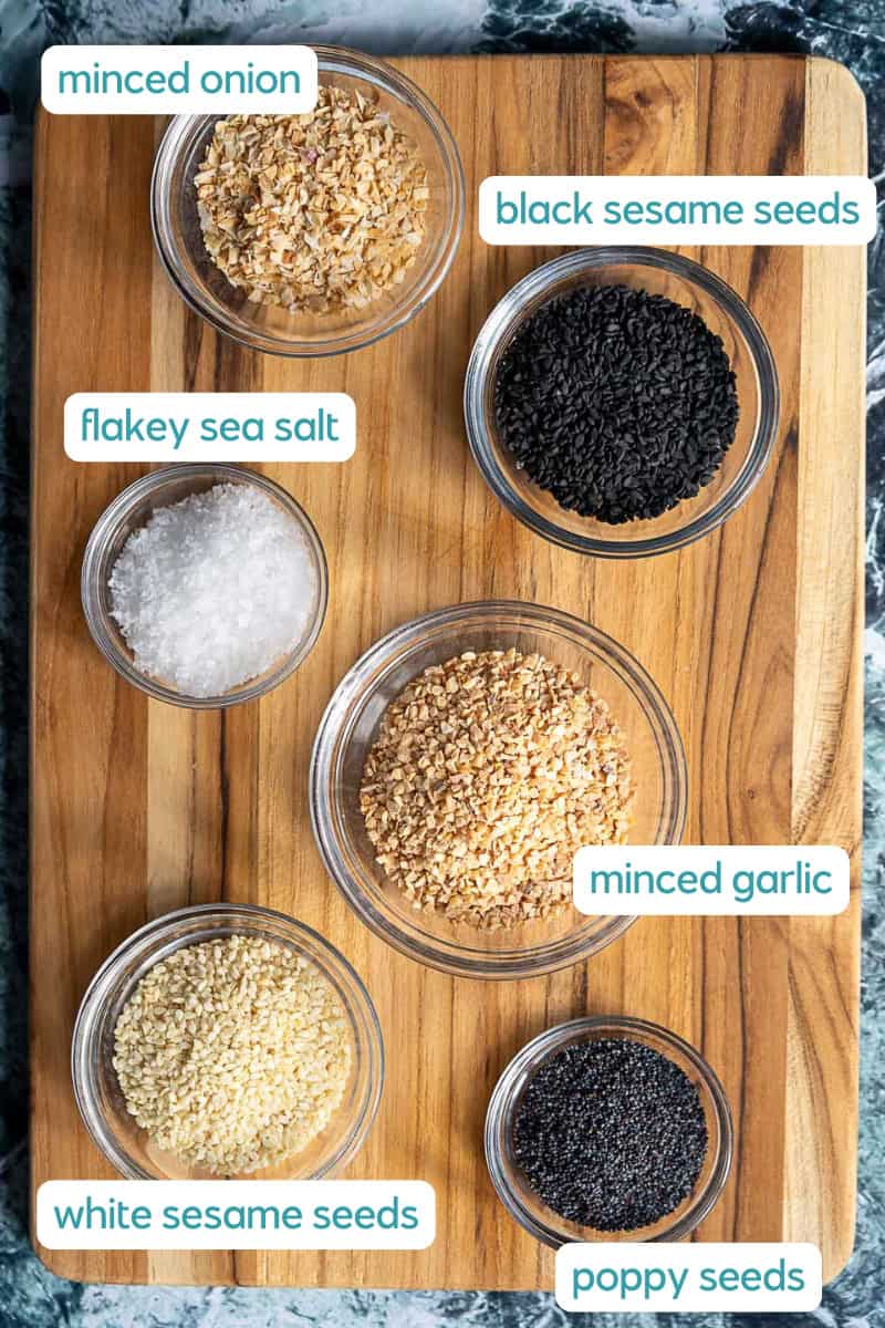 A wooden board with six small glass bowls containing labeled ingredients: minced onion, black sesame seeds, flakey sea salt, minced garlic, white sesame seeds, and poppy seeds.
