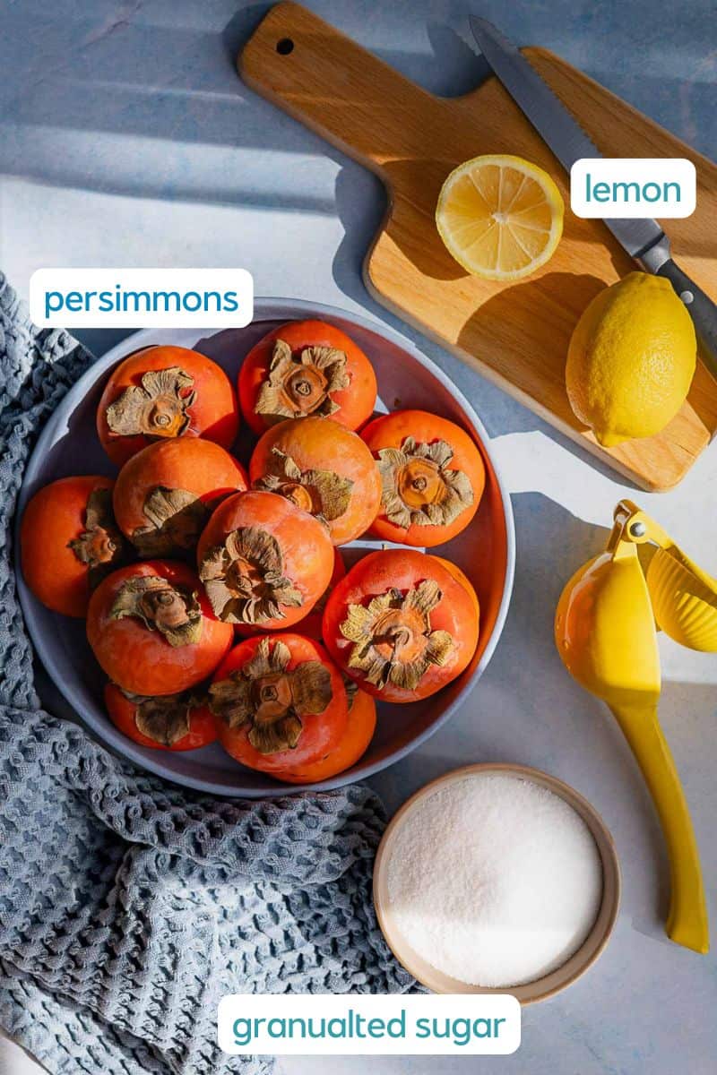 A bowl of ripe persimmons, a halved lemon with a knife on a cutting board, a lemon juicer, and granulated sugar are arranged on a light surface with a textured cloth—perfect for making homemade persimmon jam.
