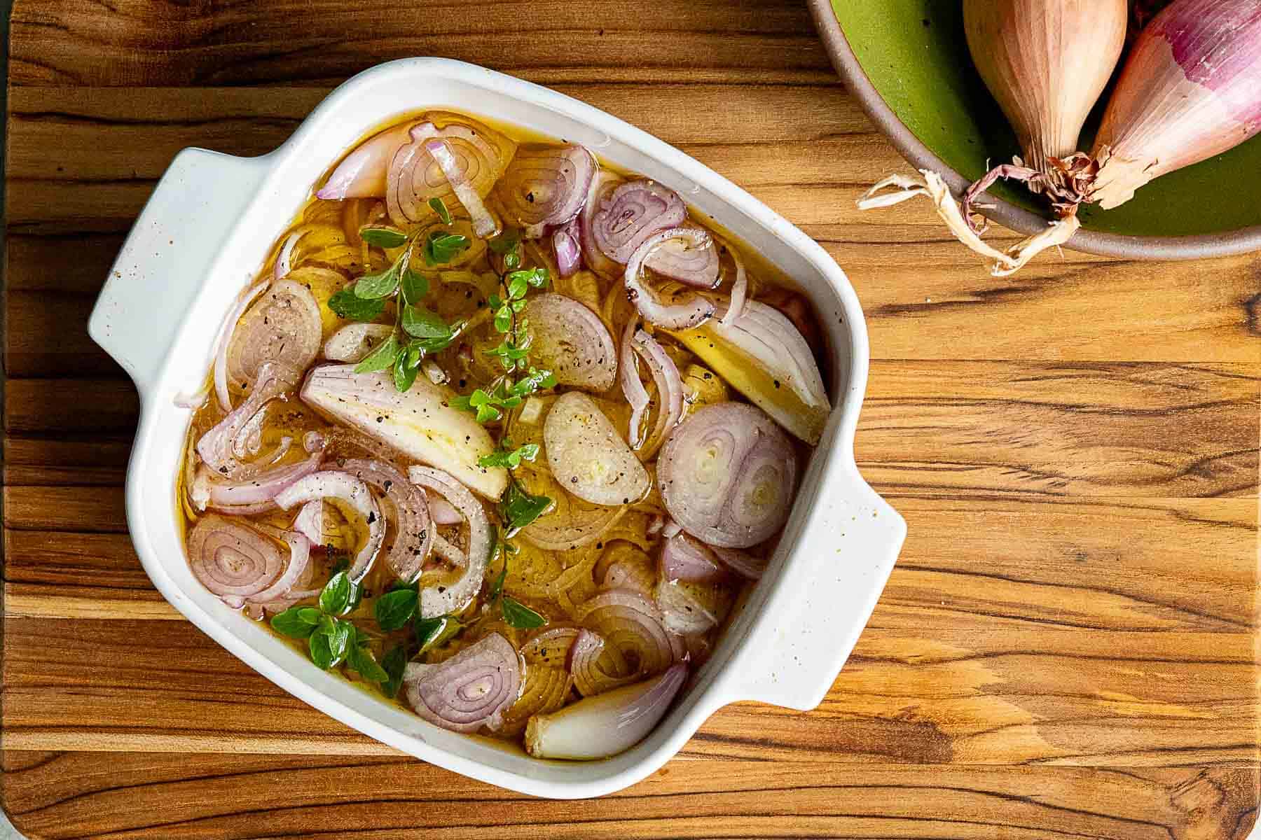 A white baking dish with sliced shallots, fresh herbs, and oil sits on a wooden surface next to a green bowl of whole shallots, ready to create complex flavor profiles in your next meal.