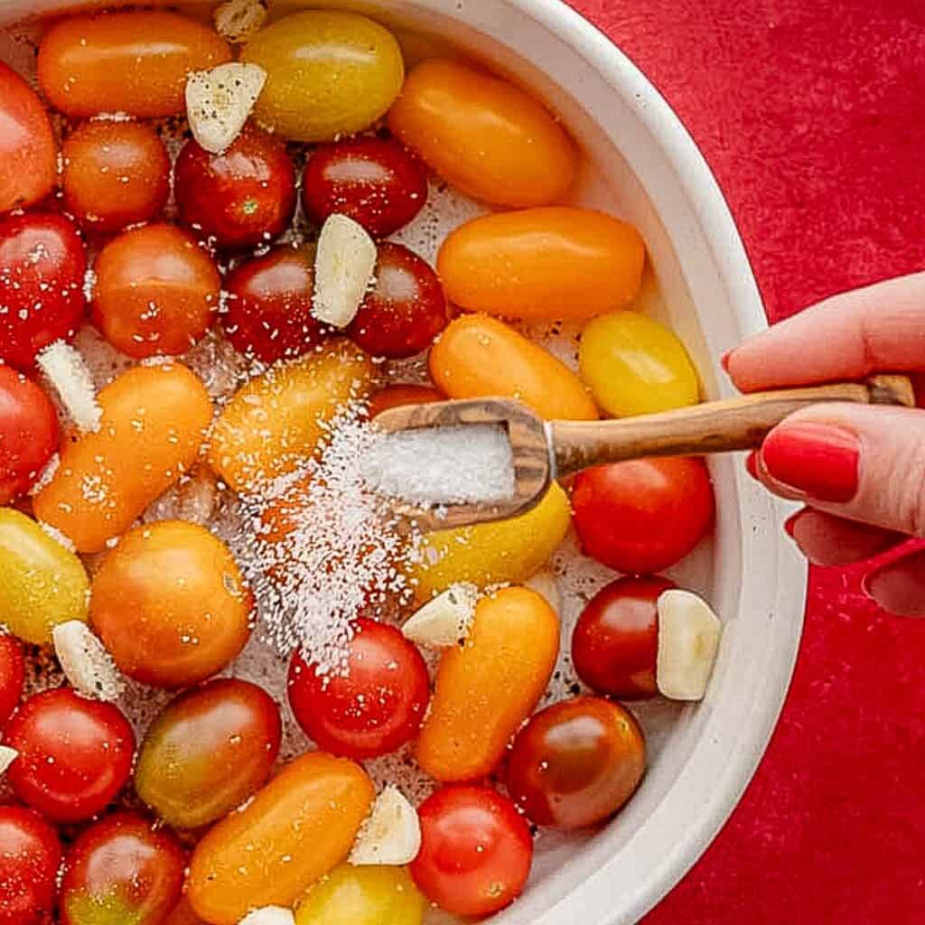 A hand with red nail polish sprinkles salt from a small wooden scoop onto a bowl of colorful cherry tomatoes and garlic cloves, enhancing their flavor profiles against a vivid red background.