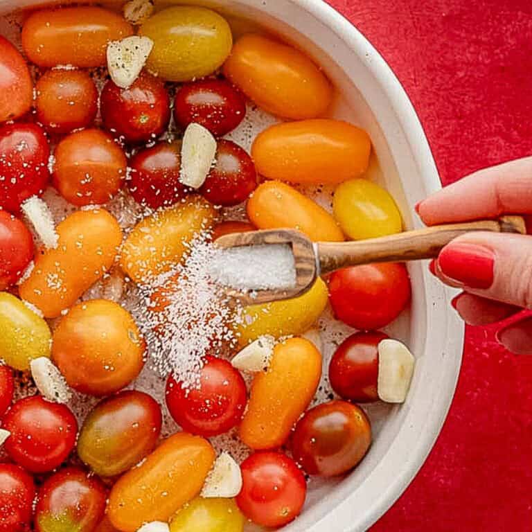 A hand with red nail polish sprinkles salt from a small wooden scoop onto a bowl of colorful cherry tomatoes and garlic cloves, enhancing their flavor profiles against a vivid red background.