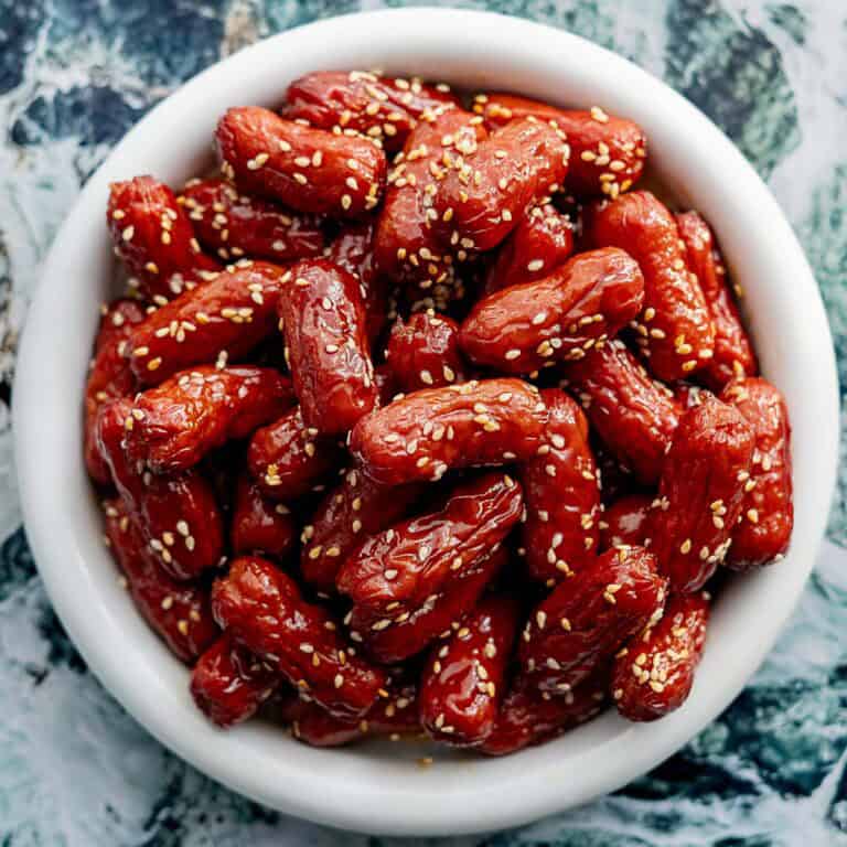 A white bowl filled with glazed cocktail sausages topped with sesame seeds, placed on a marble-patterned surface.