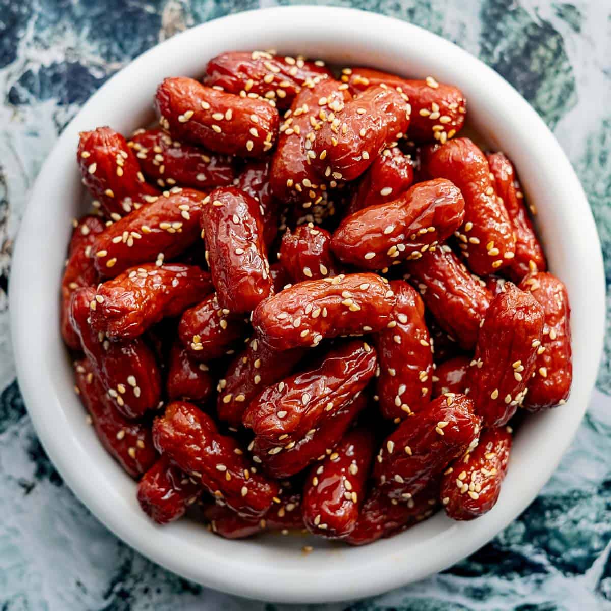 A white bowl filled with glazed cocktail sausages topped with sesame seeds, placed on a marble-patterned surface.