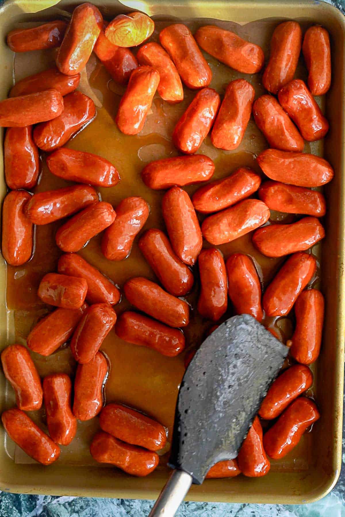 A baking sheet filled with cocktail sausages coated in sauce, with plump cocktail sausages glistening and a black spatula partially visible on the lower right side.
