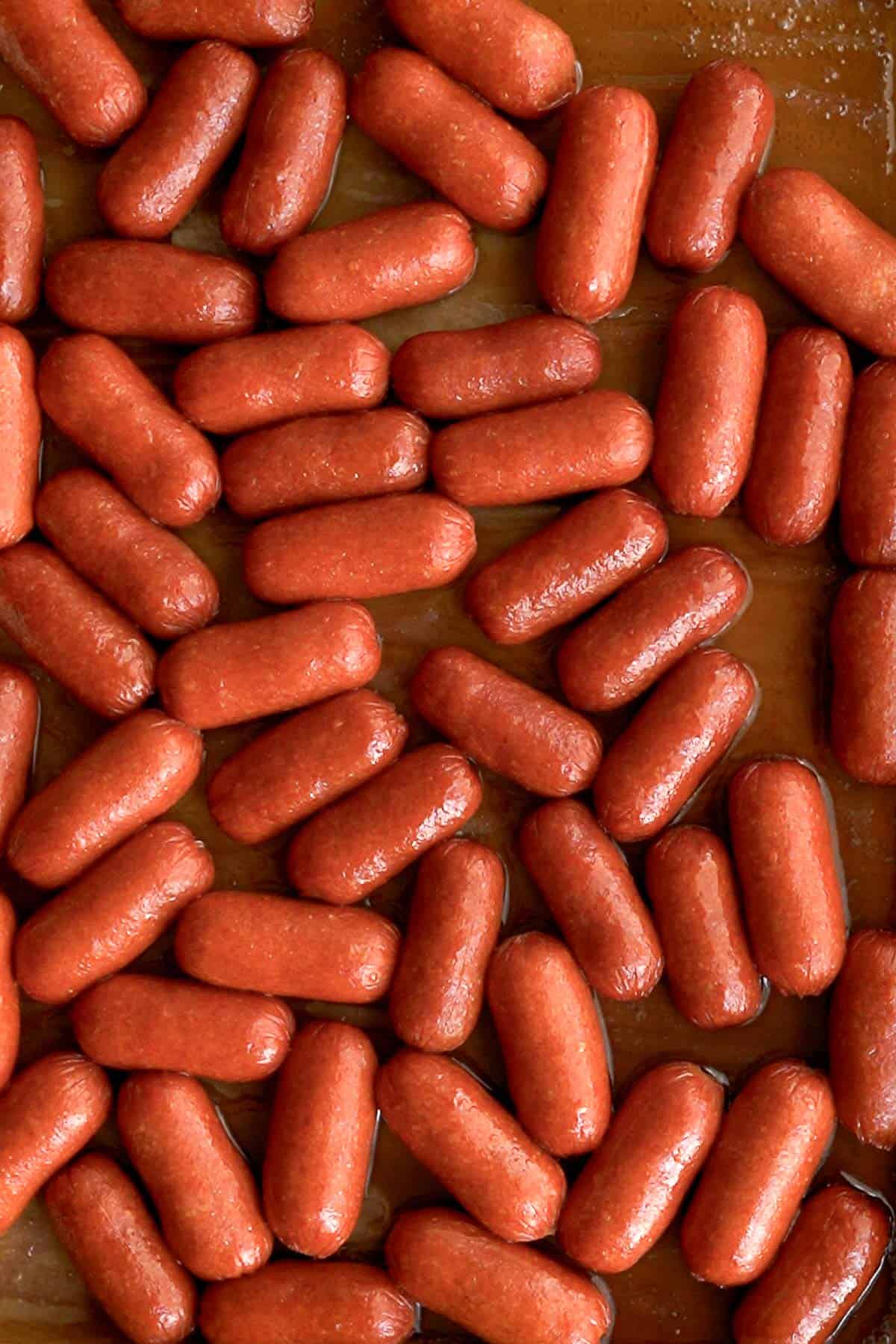 A close-up view of many cocktail sausages arranged in rows on a brown baking sheet. The shiny cocktail sausages are evenly spaced, ready to be cooked or served.