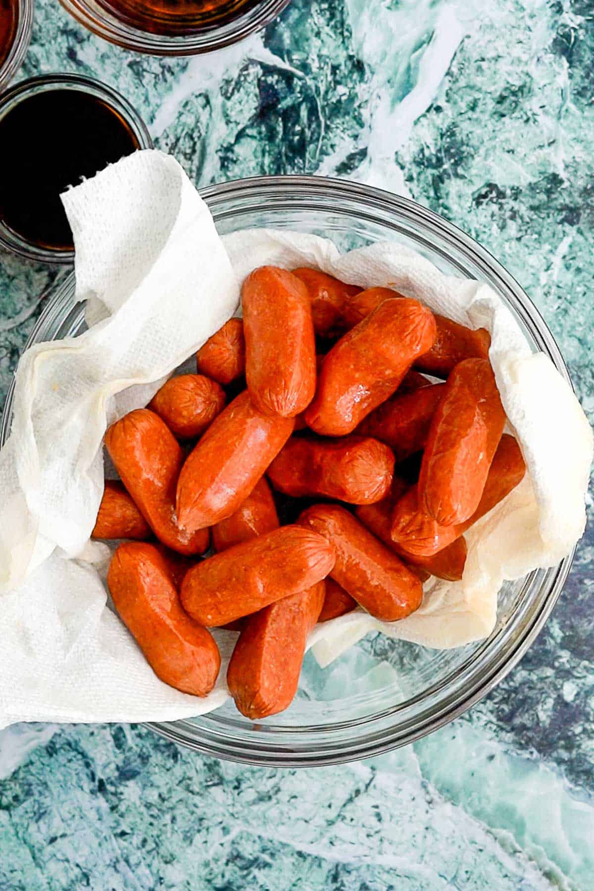 A glass bowl lined with paper towels holds several savory cocktail sausages. The bowl rests on a green and white marbled surface, surrounded by glimpses of other tempting dishes.