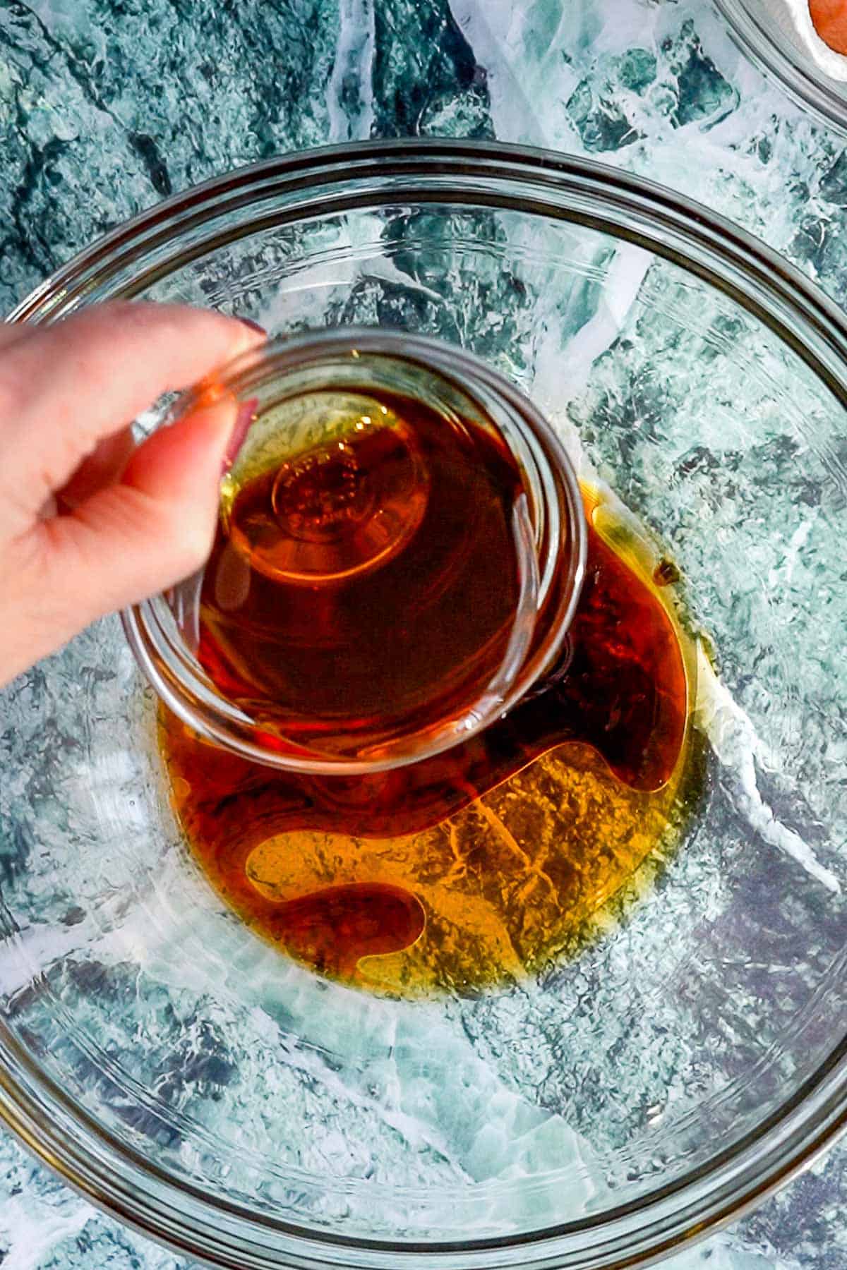 A hand pours a small glass bowl of amber liquid, likely maple syrup, into a larger glass mixing bowl on a blue and white marble surface—perfect for glazing cocktail sausages.