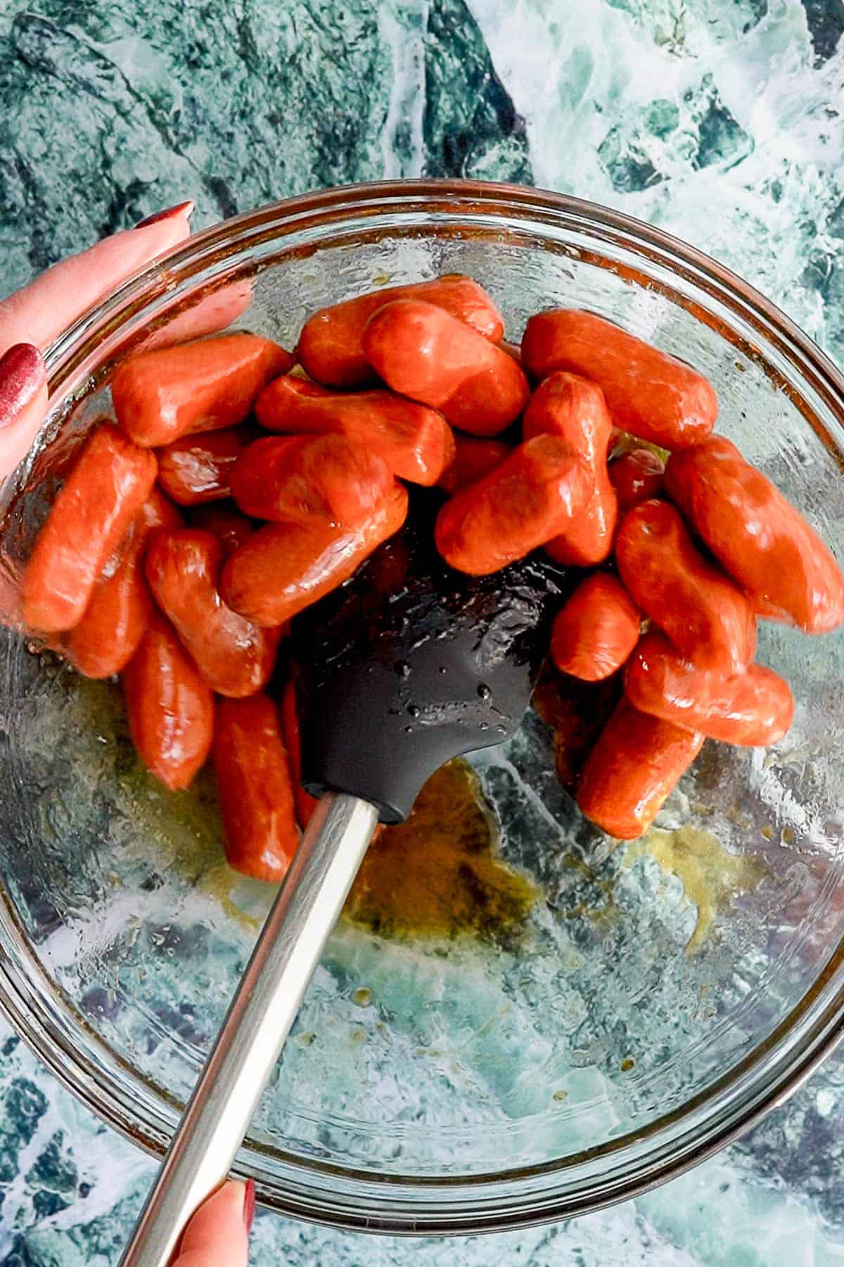 A hand holds a glass bowl filled with cocktail sausages being mixed with sauce using a black spatula, set on a green and white marbled surface—perfect for serving these flavorful cocktail sausages at your next gathering.