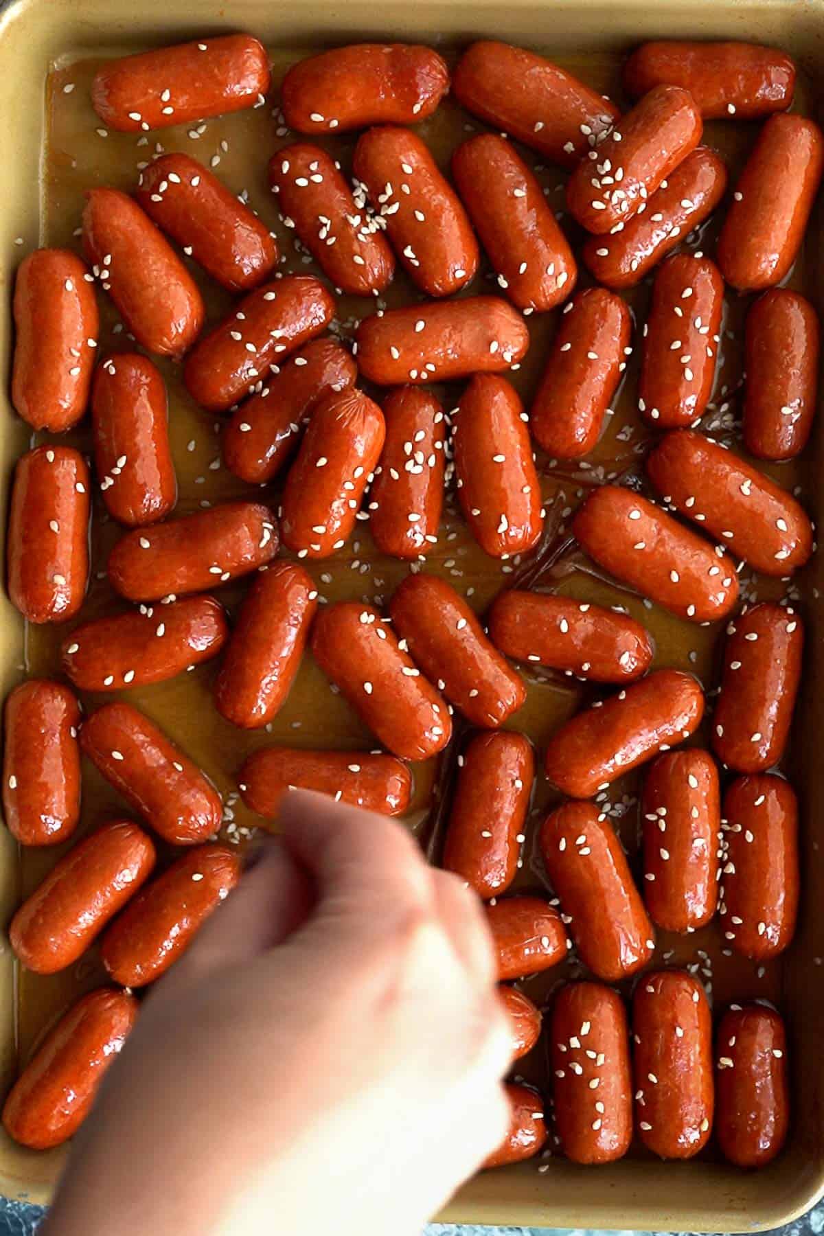 A hand sprinkles sesame seeds over a baking tray filled with shiny, glazed cocktail sausages, creating an irresistible appetizer.
