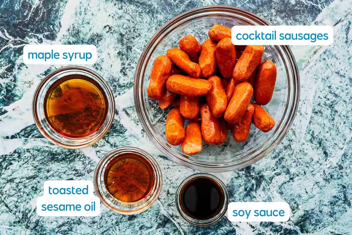 A glass bowl of cocktail sausages sits beside small bowls of maple syrup, toasted sesame oil, and soy sauce on a marble countertop. Each ingredient—including the cocktail sausages—is labeled with text.