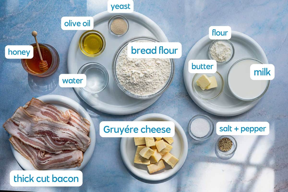 Ingredients for baking are arranged on a blue surface, including bread flour, flour, yeast, olive oil, honey, milk, butter, water, salt, pepper, thick-cut bacon, and cubed Gruyère cheese, each labeled.