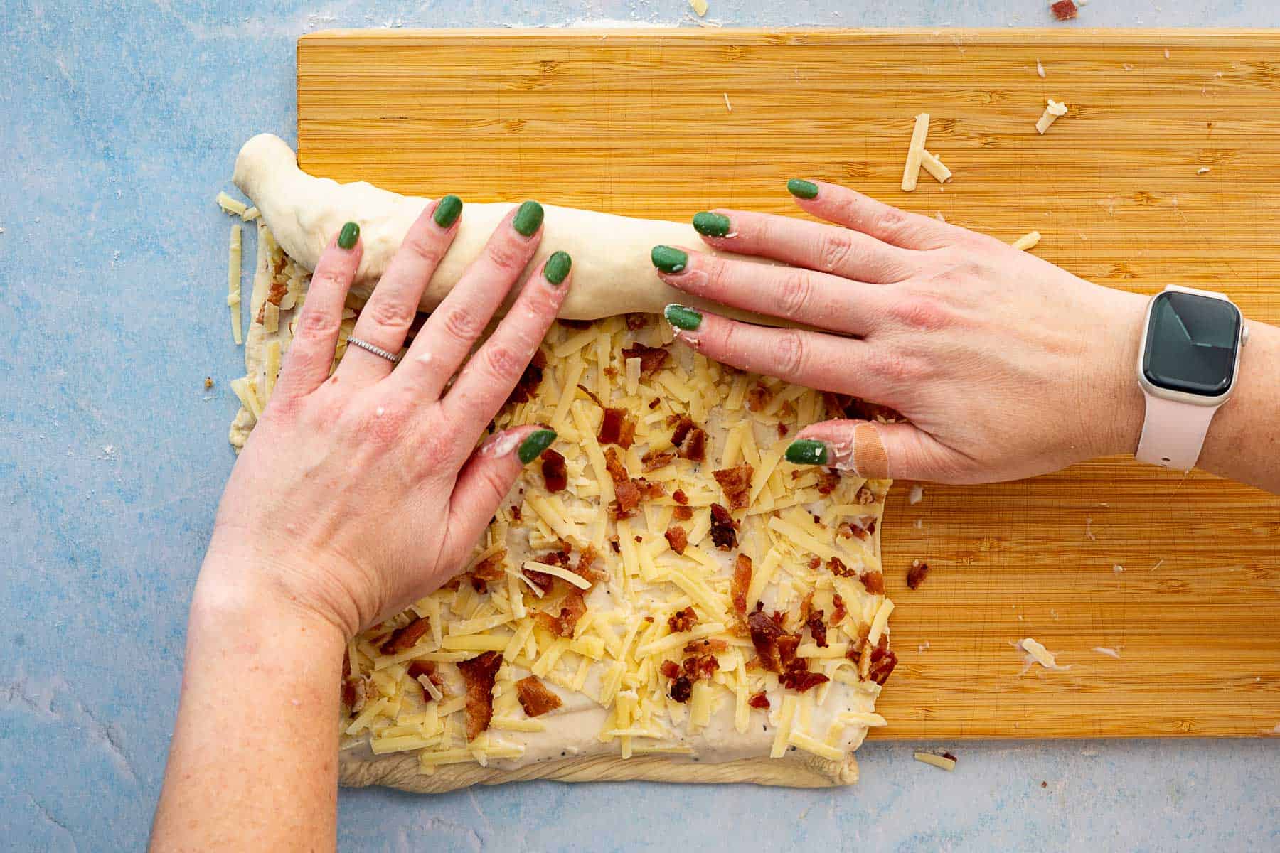 A person with green nail polish and a smartwatch is rolling up dough topped with grated cheese and bacon on a wooden cutting board.
